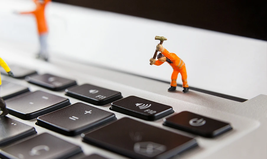 Logicminds-blog0-webmaintenance Miniature construction worker in orange overalls hammers laptop key, symbolizing tech repairs.