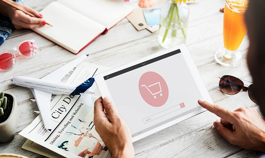 WooCommerce Marketing Tips (1) Person uses tablet with shopping cart icon, surrounded by travel magazines, airplane, and drinks.